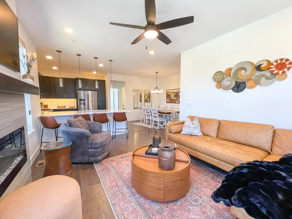 A cozy, open-concept living and dining area featuring a leather sofa, round coffee table, artistic wall decor, a modern fireplace, and a visible dining area. The space is bathed in warm lighting, inviting guests to gather, relax, and share stories by the fire or around the table.