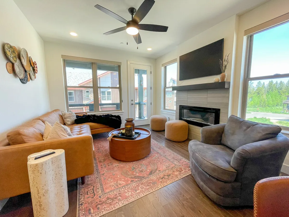 The image showcases a warm, inviting living space with a plush leather sofa, stylish coffee table, and a glowing fireplace beneath tasteful artwork. Sunlight streams through expansive windows, bathing the room in natural light and offering breathtaking mountain views, while the cozy setup invites guests to relax, socialize, or enjoy a quiet evening as the sun sets.