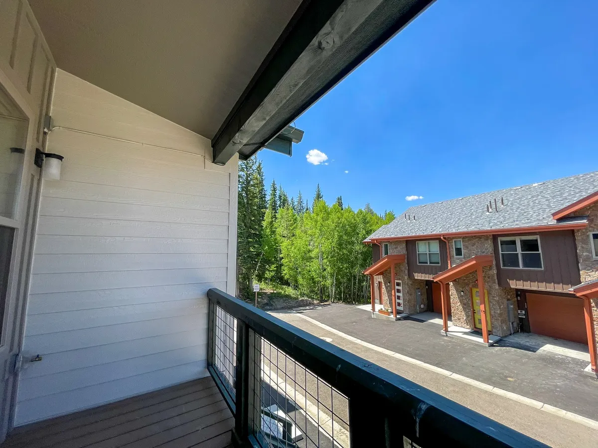 An exterior shot from a balcony, featuring a sturdy railing, surrounding trees, and glimpses of adjacent buildings. The setting is peaceful and encourages leisurely activities like enjoying coffee or simply soaking in the views.