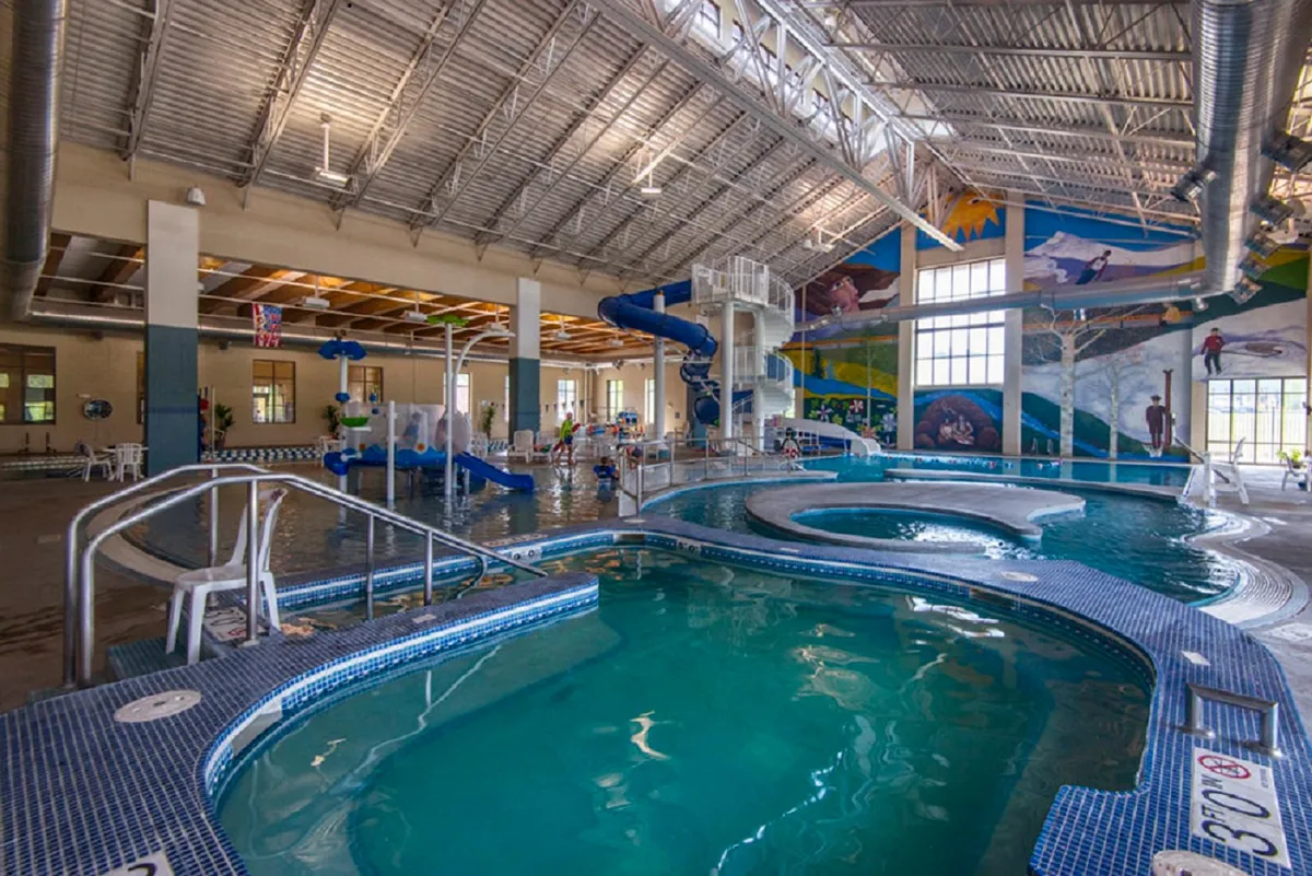 Inside a recreation center, children and families are seen splashing and playing on waterslides, in a large pool area adorned with playful murals and vibrant structures. The lively atmosphere is filled with laughter and activity, making it an inviting spot for all-weather fun.