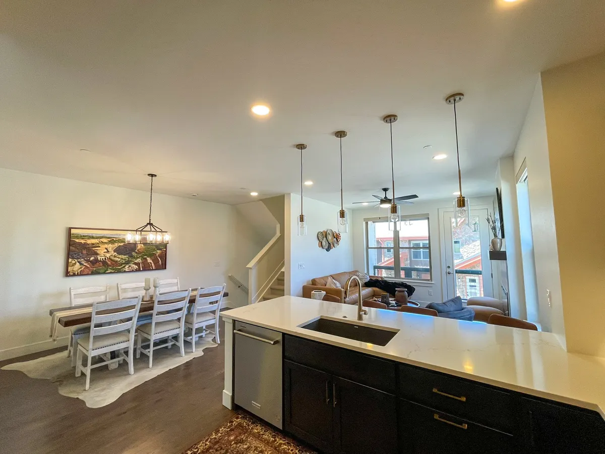 A bright, open living, dining, and kitchen area featuring a large dining table, kitchen island, plush sofa, pendant lighting, and expansive windows that connect to the outdoor patio. The space exudes a welcoming atmosphere with modern amenities and a view to the outdoors.