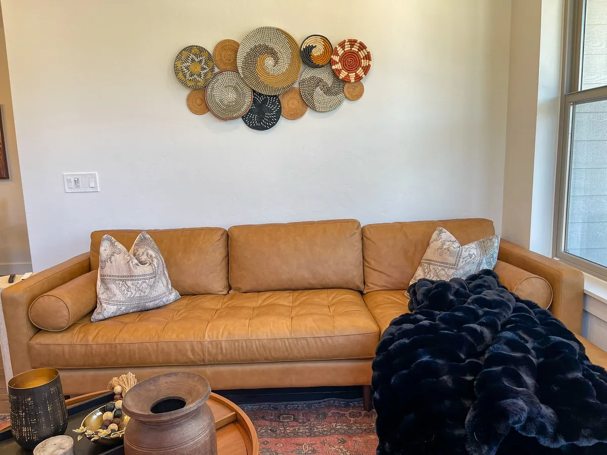 A cozy living area with a leather couch, decorative pillows, woven wall art, a coffee table, and a textured blanket. The space is ideal for relaxing, socializing, or enjoying a movie night.