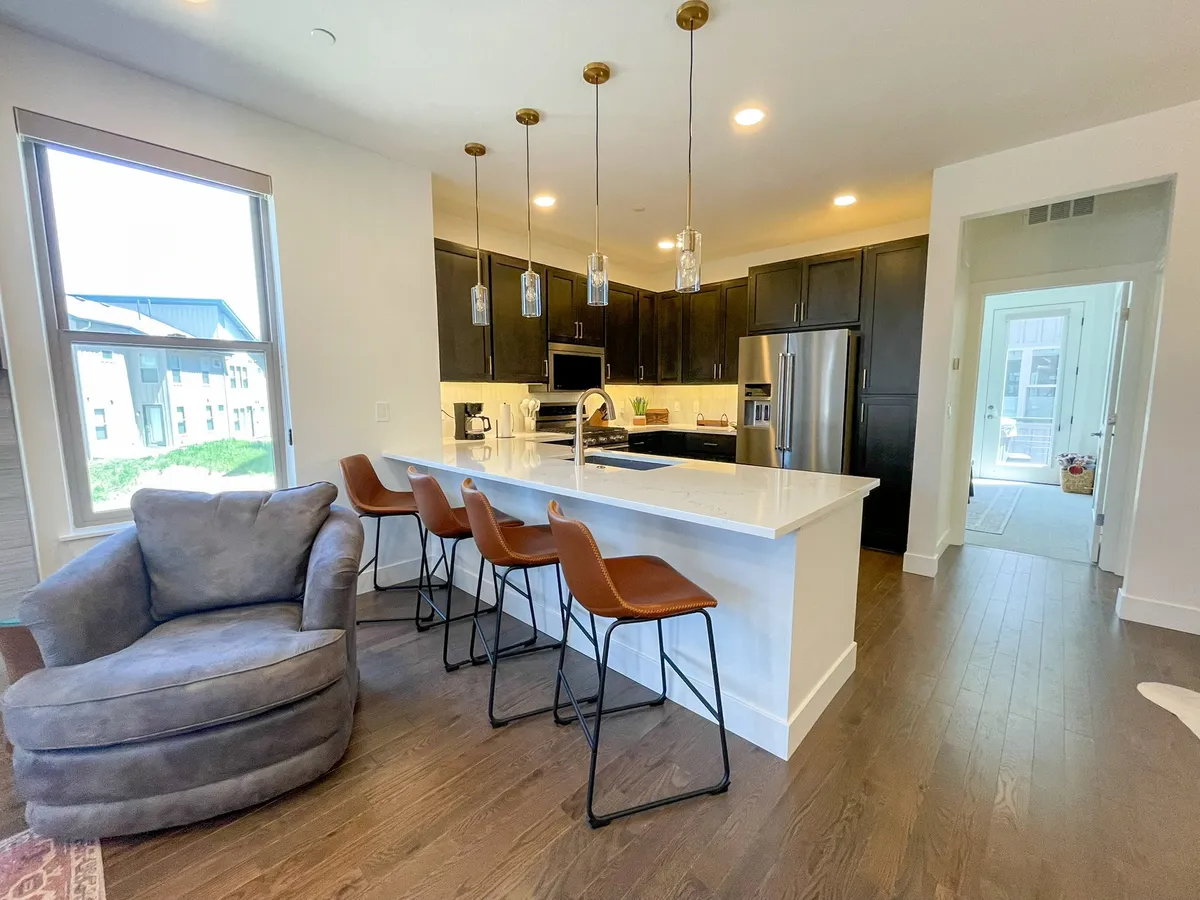A stylish kitchen interior with a spacious island, bar stools, elegant pendant lighting, and a window view. The layout invites socializing, cooking, and entertaining, evoking feelings of warmth and sophistication.