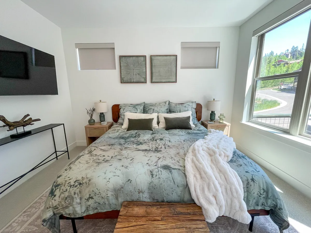 A tranquil bedroom with a plush bed adorned in floral bedding, a wooden bench at the foot, decorative wall art, and a smart TV mounted on the wall. Sunlight streams through the window, creating a warm, calming atmosphere ideal for rest and relaxation.