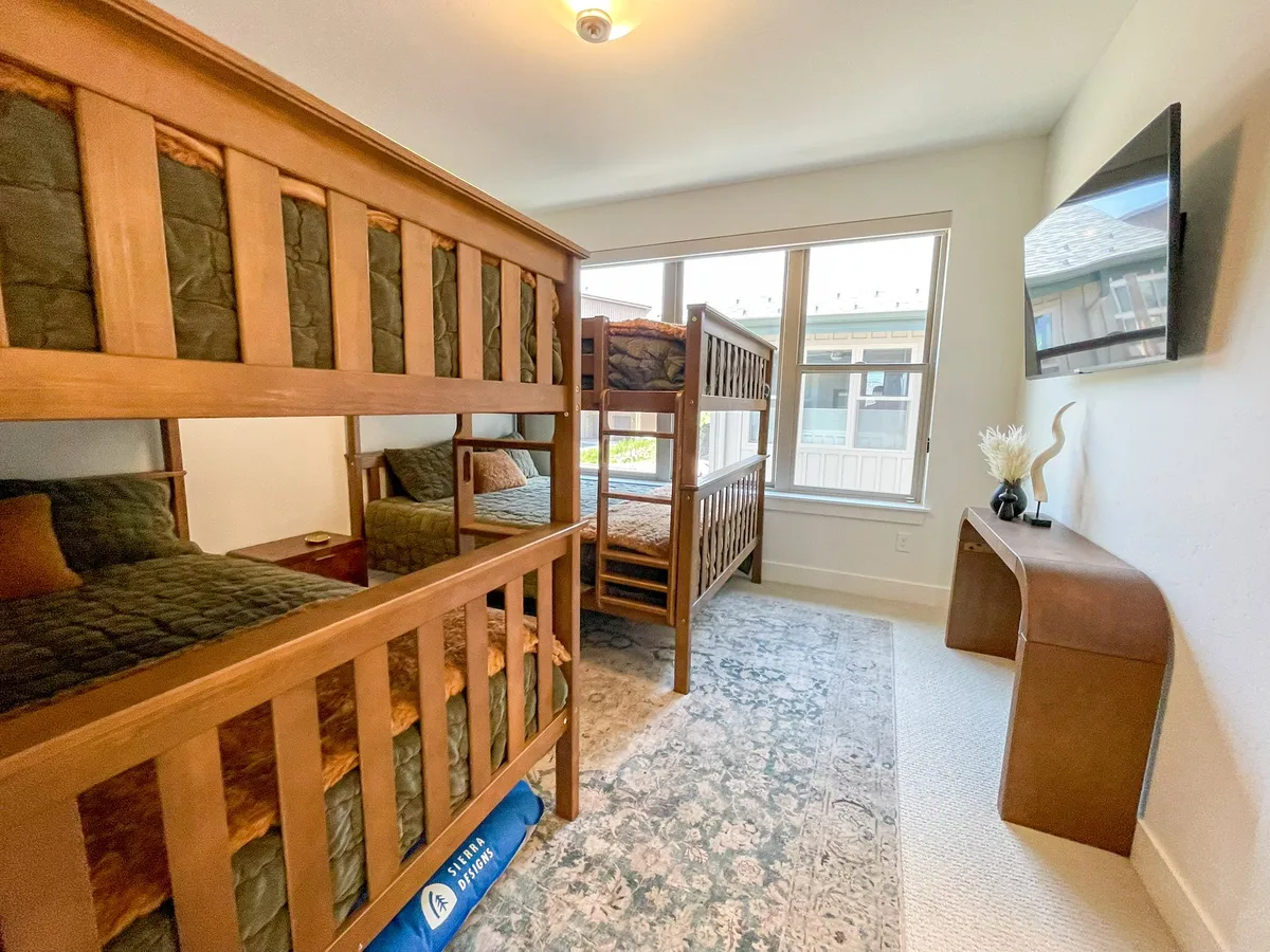 A spacious bunk room featuring custom bunk beds with queen-over-queen and full-over-full arrangements, a rug, a television, and natural light pouring in from windows. The room exudes a family-friendly, playful vibe, perfect for children or groups.