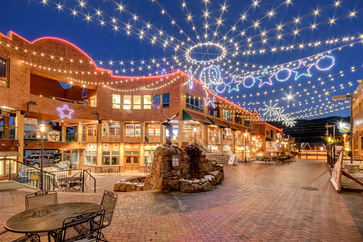A lively exterior scene in downtown Winter Park featuring string lights overhead, decorative stars, a stone fountain, inviting outdoor seating, and storefronts. People can be seen dining, shopping, strolling, and socializing in a festive atmosphere.